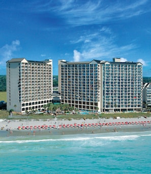 Un hotel frente al mar con varios pisos, piscina y sombrillas de playa.
