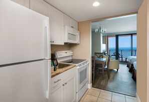 Una cocina con armarios blancos, microondas y estufa. Hay una zona de comedor con mesa y sillas, y un balcón con vista al océano.
