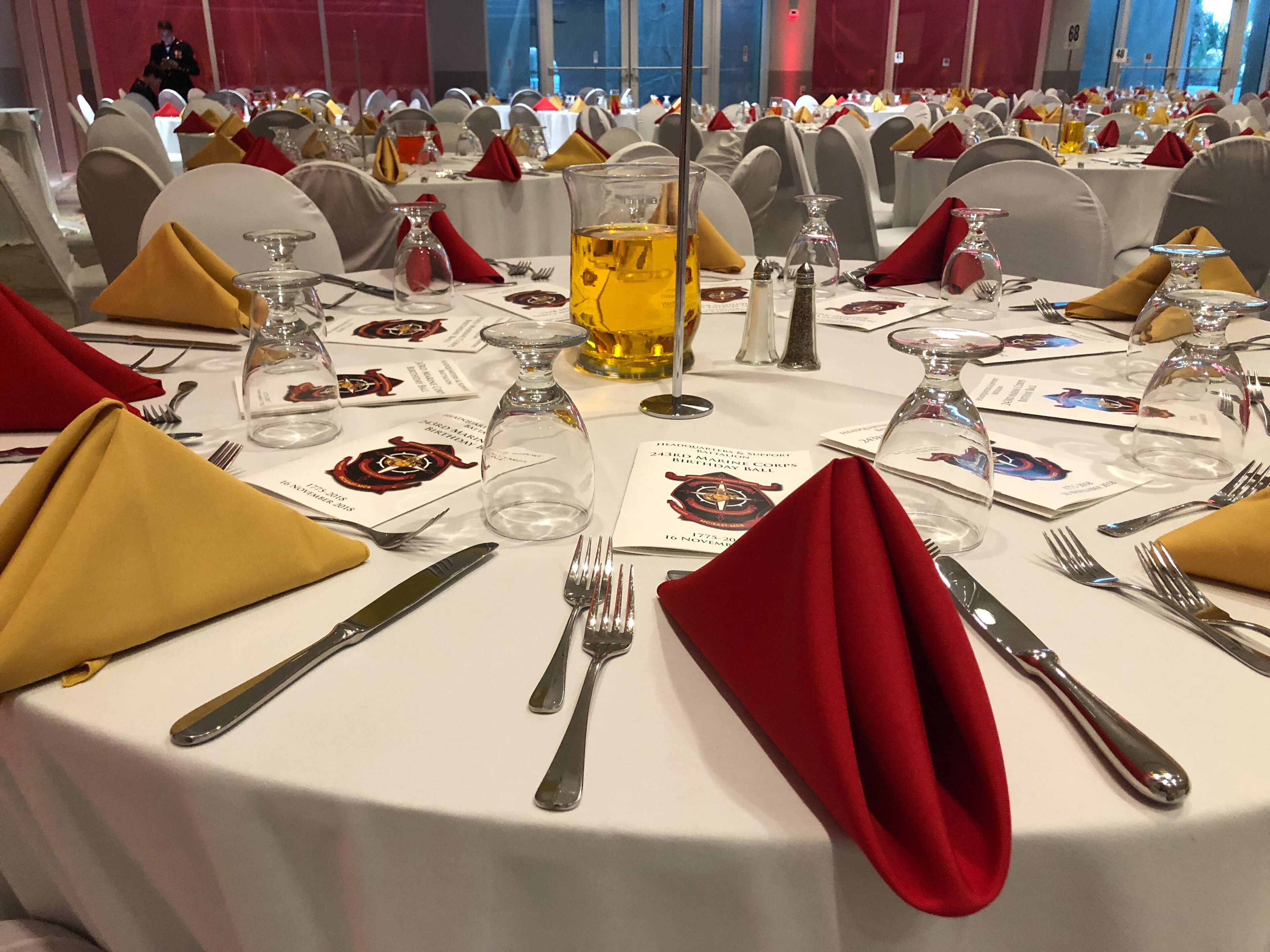 A banquet hall with round tables set for a formal event, featuring white tablecloths, red napkins, and clear glassware.