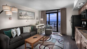 A modern living room with a grey sofa, a wooden coffee table, and a dining area with a small table and chairs. A painting of a beach scene hangs on the wall, and a large window offers a view of the ocean.