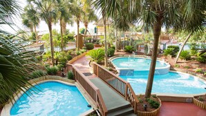 A pool area with multiple pools, wooden deck, and surrounding palm trees. - Oceanfront 1 Bedroom Suite w/ Gorgeous View + Official On-Site Rental Privileges (North Myrtle Beach)