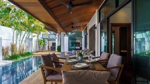 A dining area with a wooden table set for four, surrounded by cushioned chairs, overlooking a swimming pool and lush greenery. - Emotion2 - Brand New 3BR Pool Villa (Tambon Rawai)