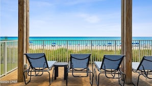 Eine hölzerne Pergola mit vier Stühlen und einem kleinen Tisch, mit Blick auf einen Strand unter wolkenlosem, blauem Himmel.