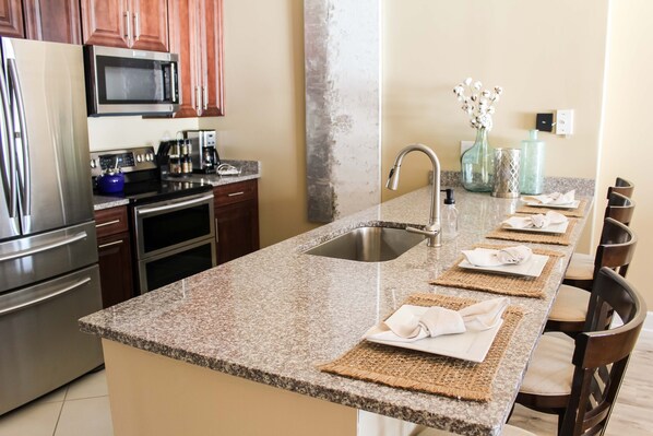 A kitchen with granite countertops, stainless steel appliances, and a long dining table set with placemats and napkins. - Beachfront Condo 22nd Penthouse Floor with 10-foot ceiling (Panama City Beach)