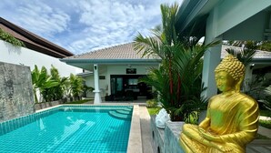 Una piscina con una estatua dorada de Buda a su lado, un edificio con techo de tejas y exuberante vegetación al fondo.
