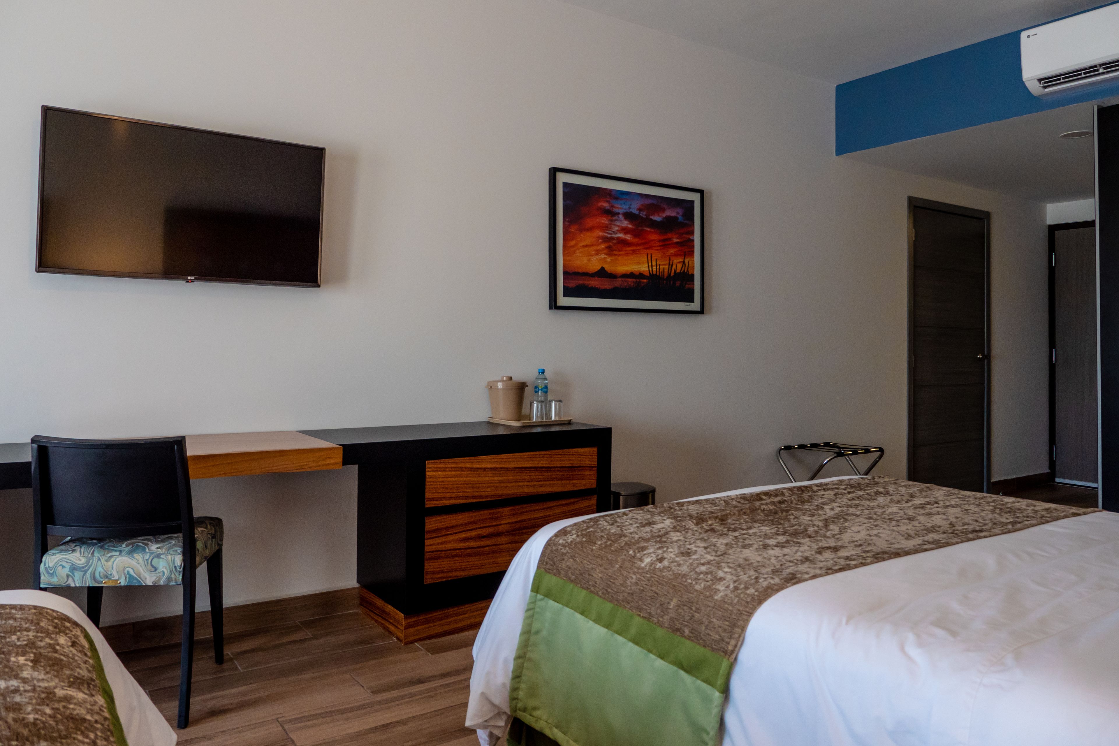 A hotel room with a bed, a desk, a chair, a TV, and a painting on the wall.