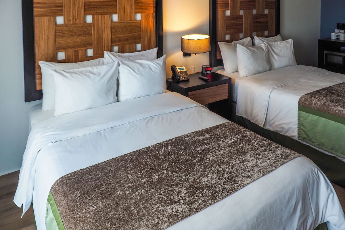 A hotel room with two beds, each with white bedding and a brown throw blanket. There is a nightstand with a lamp and a phone between the beds.