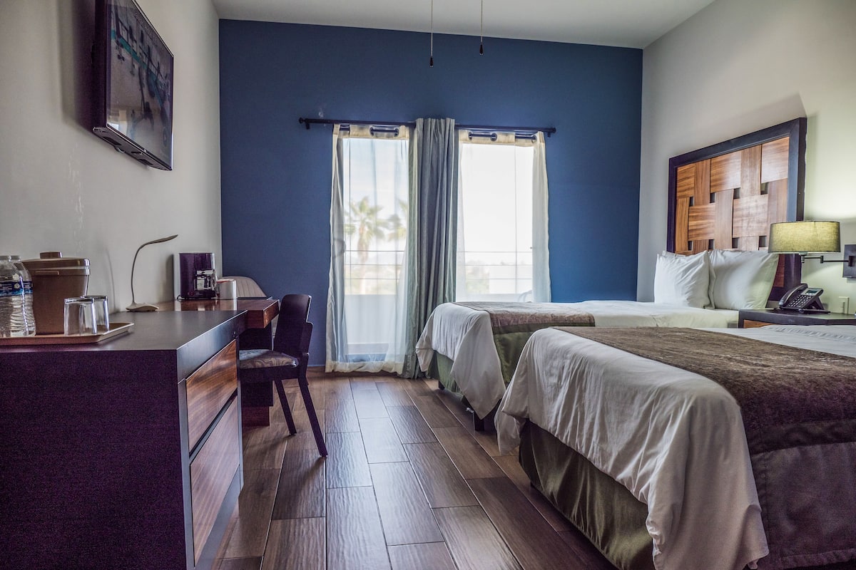 A hotel room with a desk, two beds, a window with curtains, a lamp, and a phone on the nightstand.