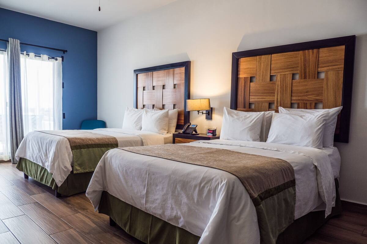 A hotel room with two beds, a nightstand with a lamp, and a blue wall.