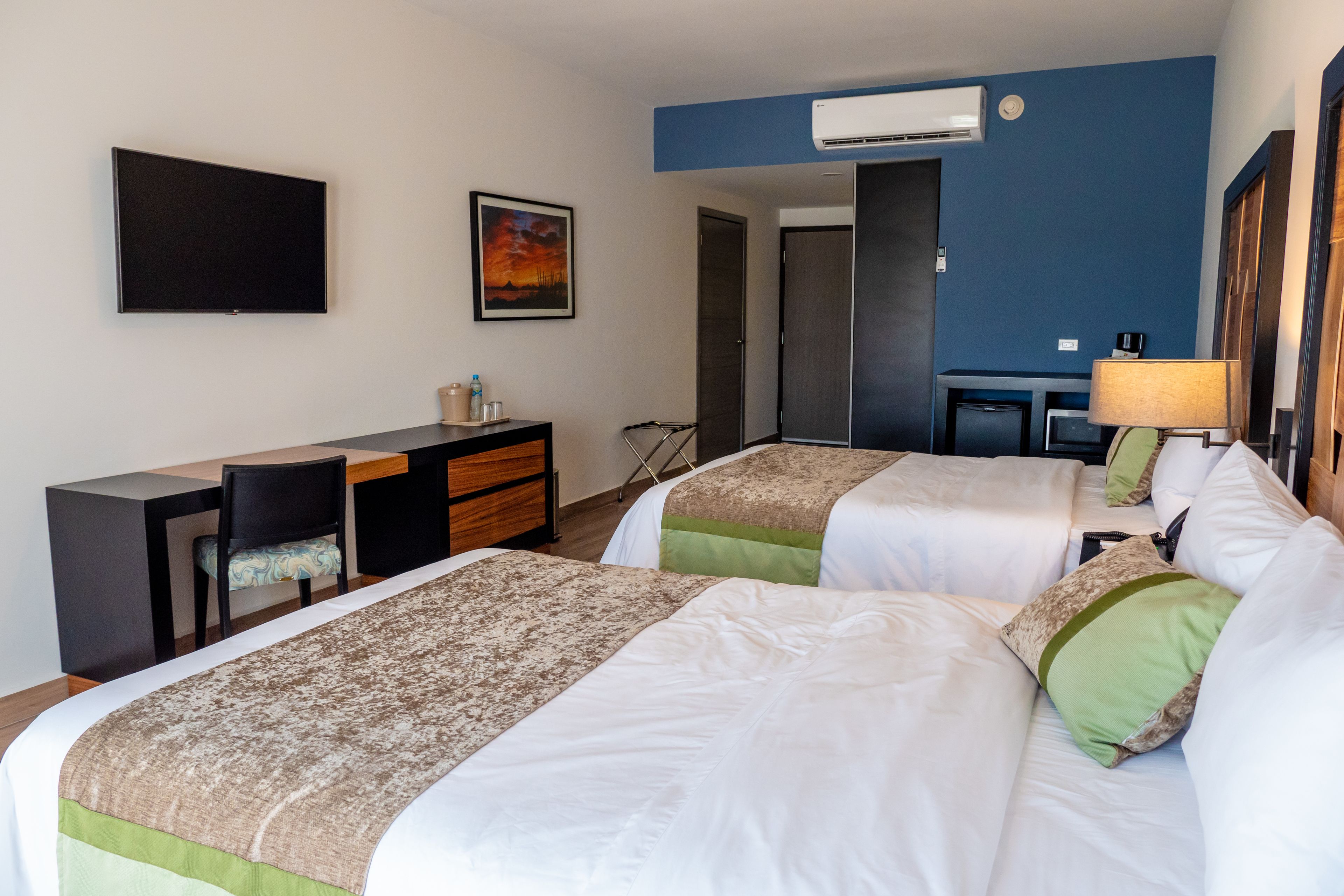 A hotel room with two beds, a desk, a chair, a TV, and an air conditioning unit.