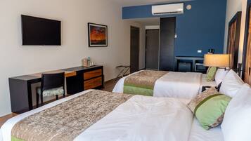 A hotel room with two beds, a desk, a chair, a TV, and an air conditioning unit.
