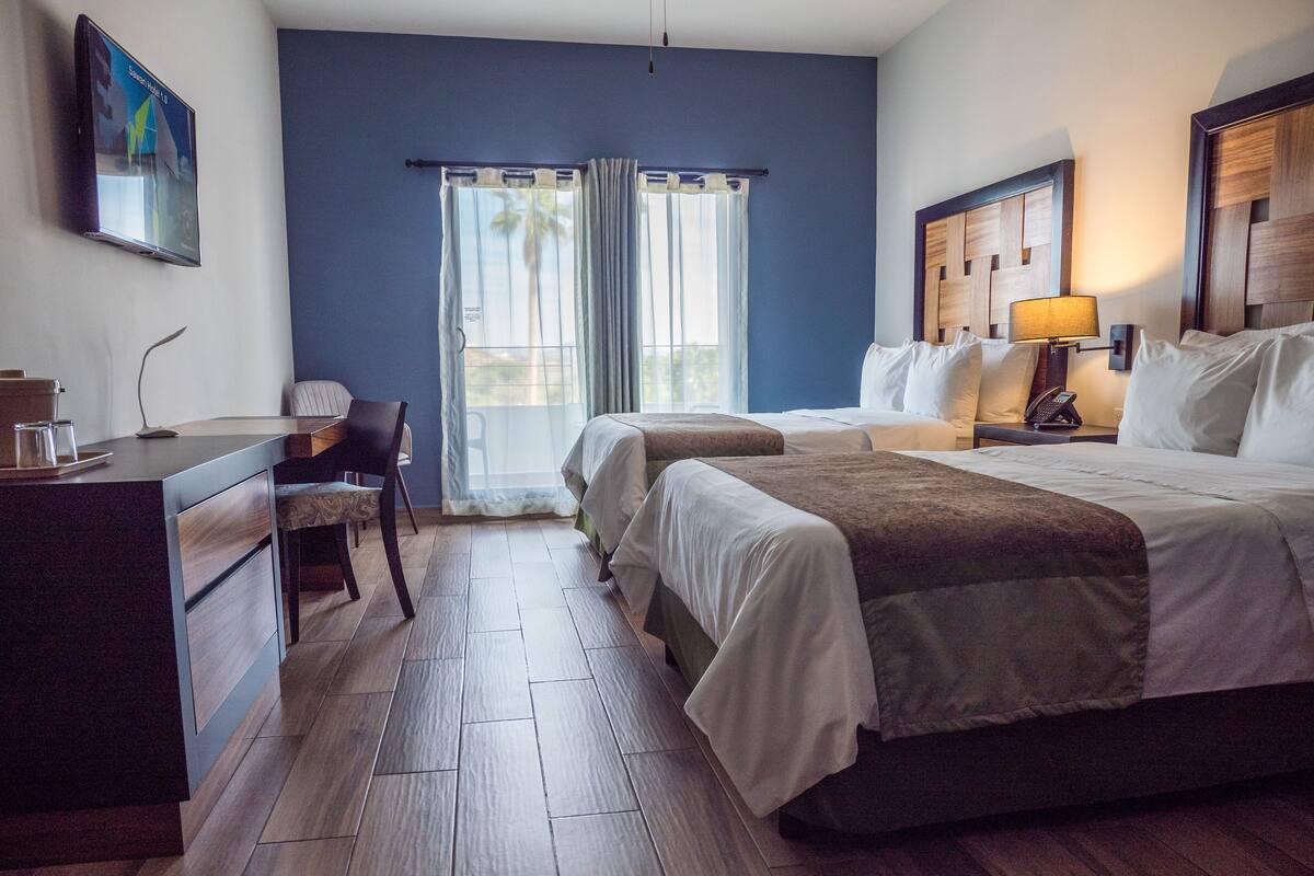 A hotel room with two beds, a desk, a chair, a nightstand with a lamp, and a window with curtains.