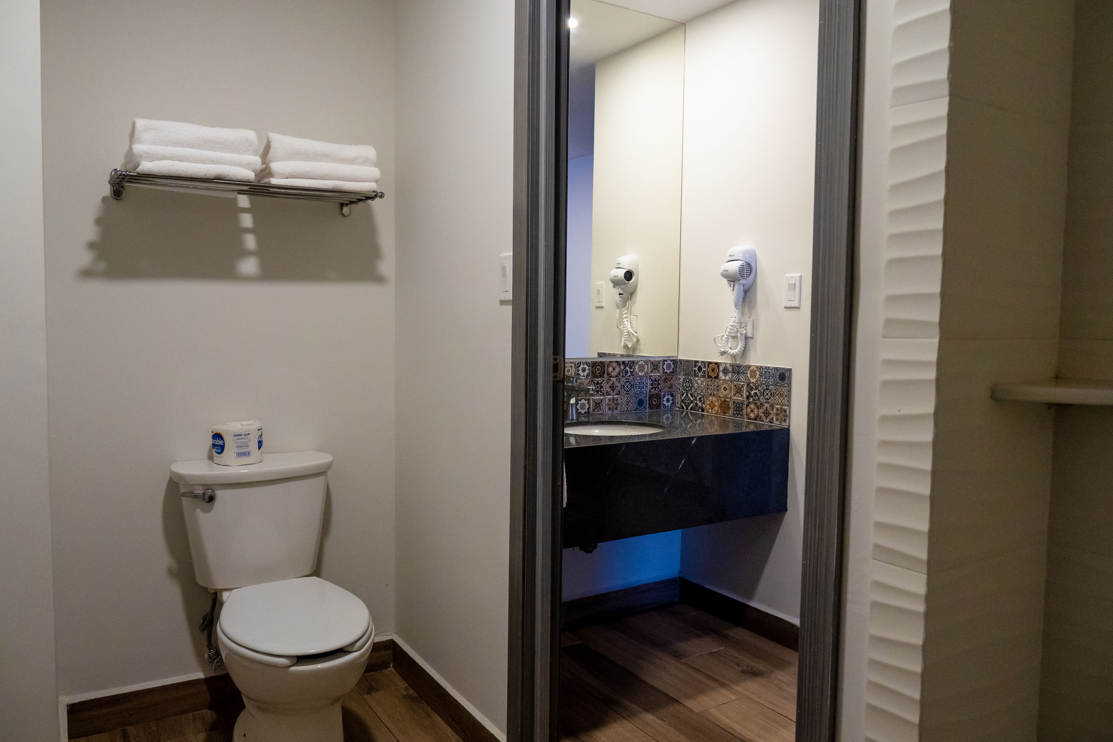 Un baño con inodoro, toallero con dos toallas, lavamanos con espejo y secador de pelo.