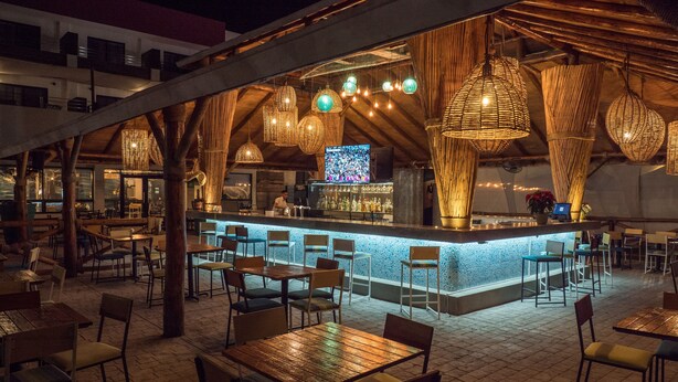 An outdoor dining area with wooden tables and chairs, a bar with a counter, and hanging wicker lamps. - Sawari Hotel (San Carlos)