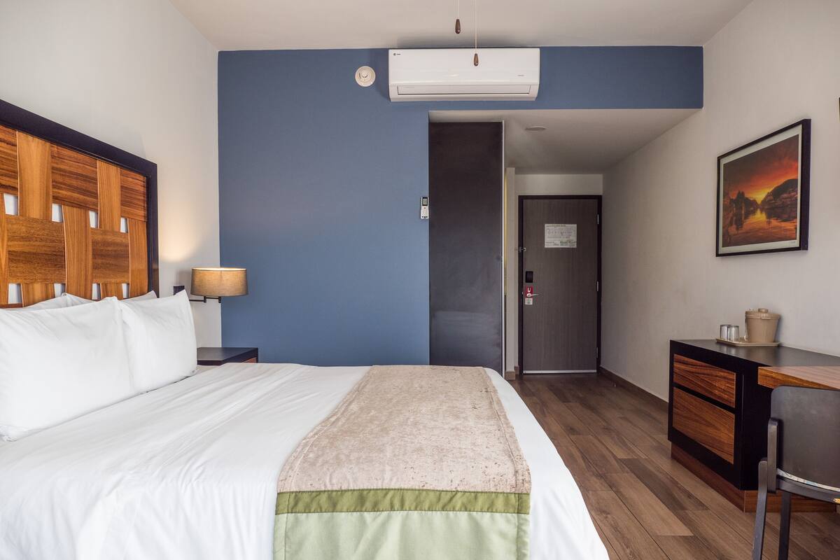 A hotel room with a bed, a desk, a chair, an air conditioning unit, and a painting on the wall.