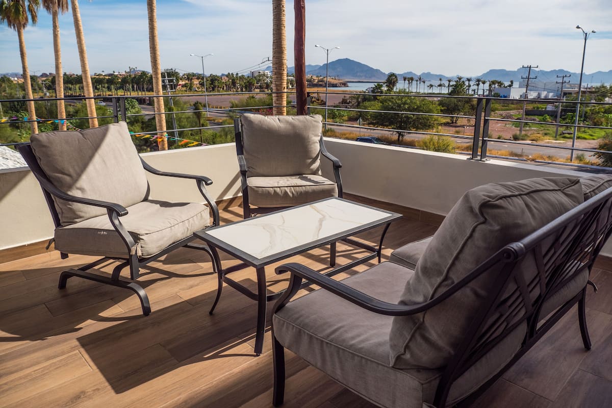 A balcony with two chairs and a table, overlooking a scenic landscape with palm trees and distant mountains.