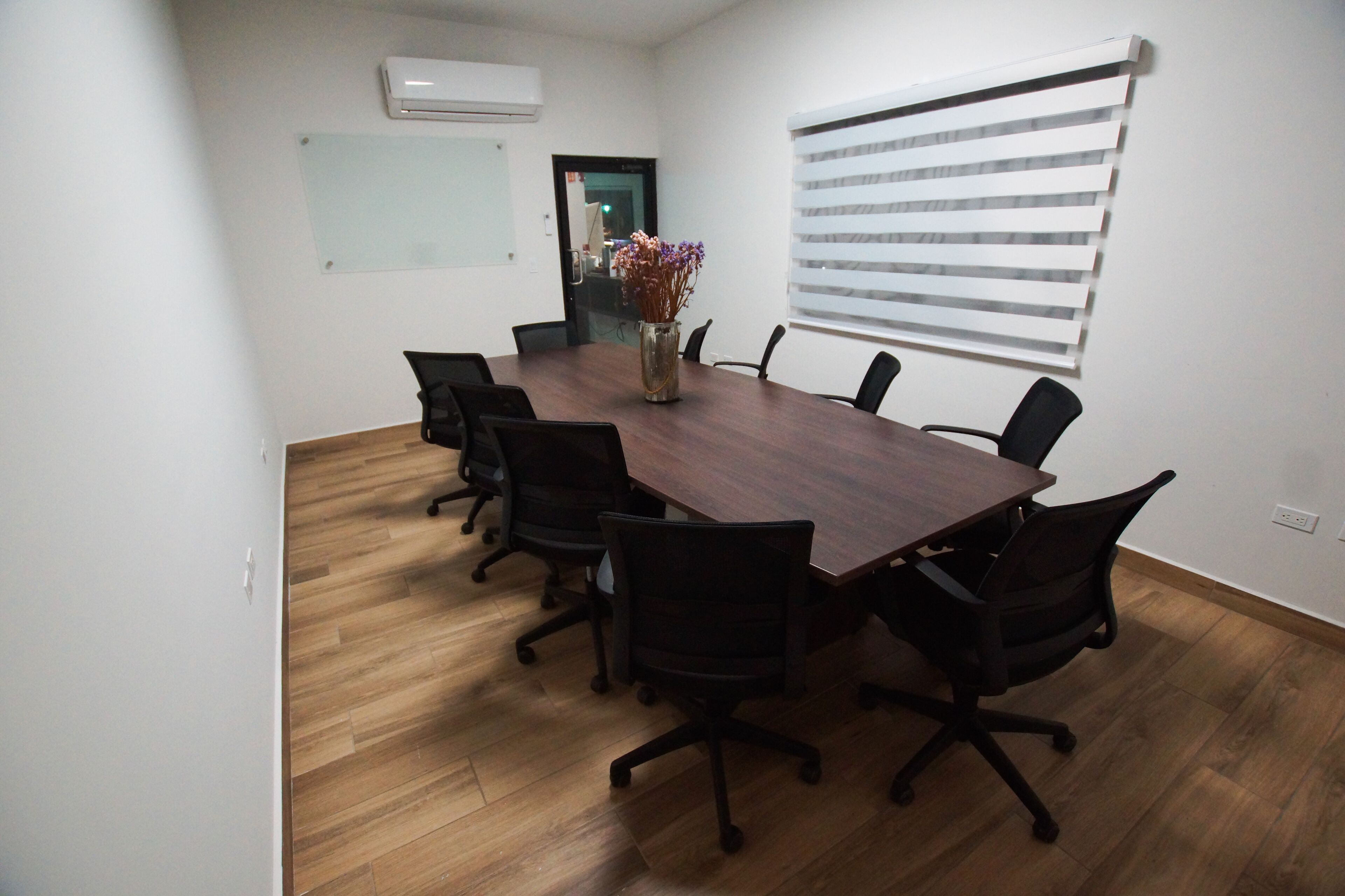 A conference room with a wooden table, black office chairs, a vase with flowers, a whiteboard, and a window with blinds.