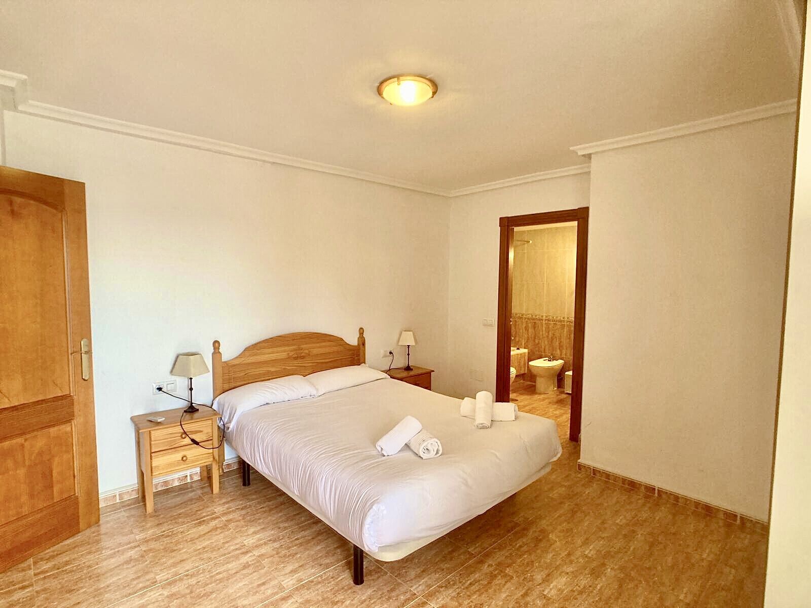 Een slaapkamer met een houten bed, nachtkastjes, een spiegel en een zichtbare badkamer door een open deur.