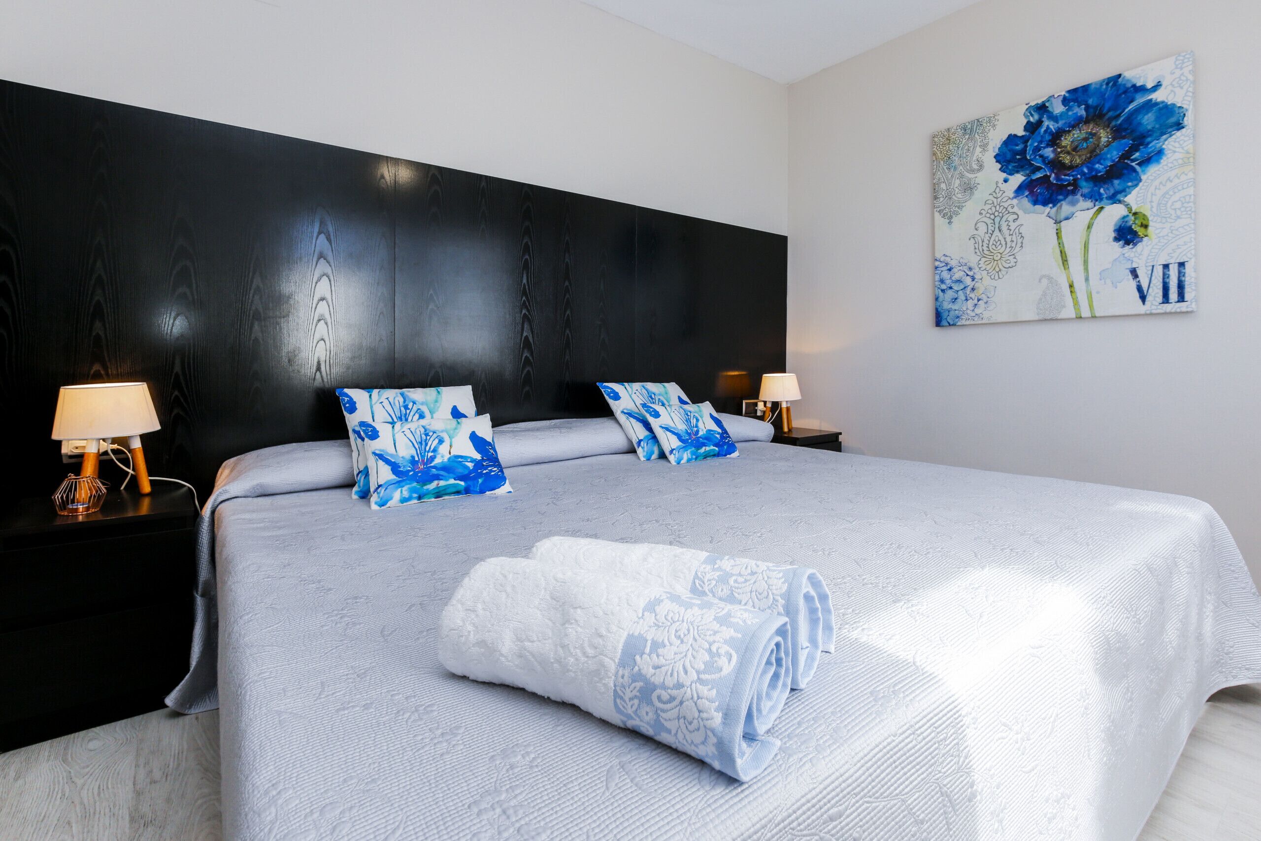 A neatly made bed with a blue and white patterned bedspread, two matching pillows, and a rolled towel on the bed. There are two bedside tables with lamps, and a large floral artwork is on the wall.