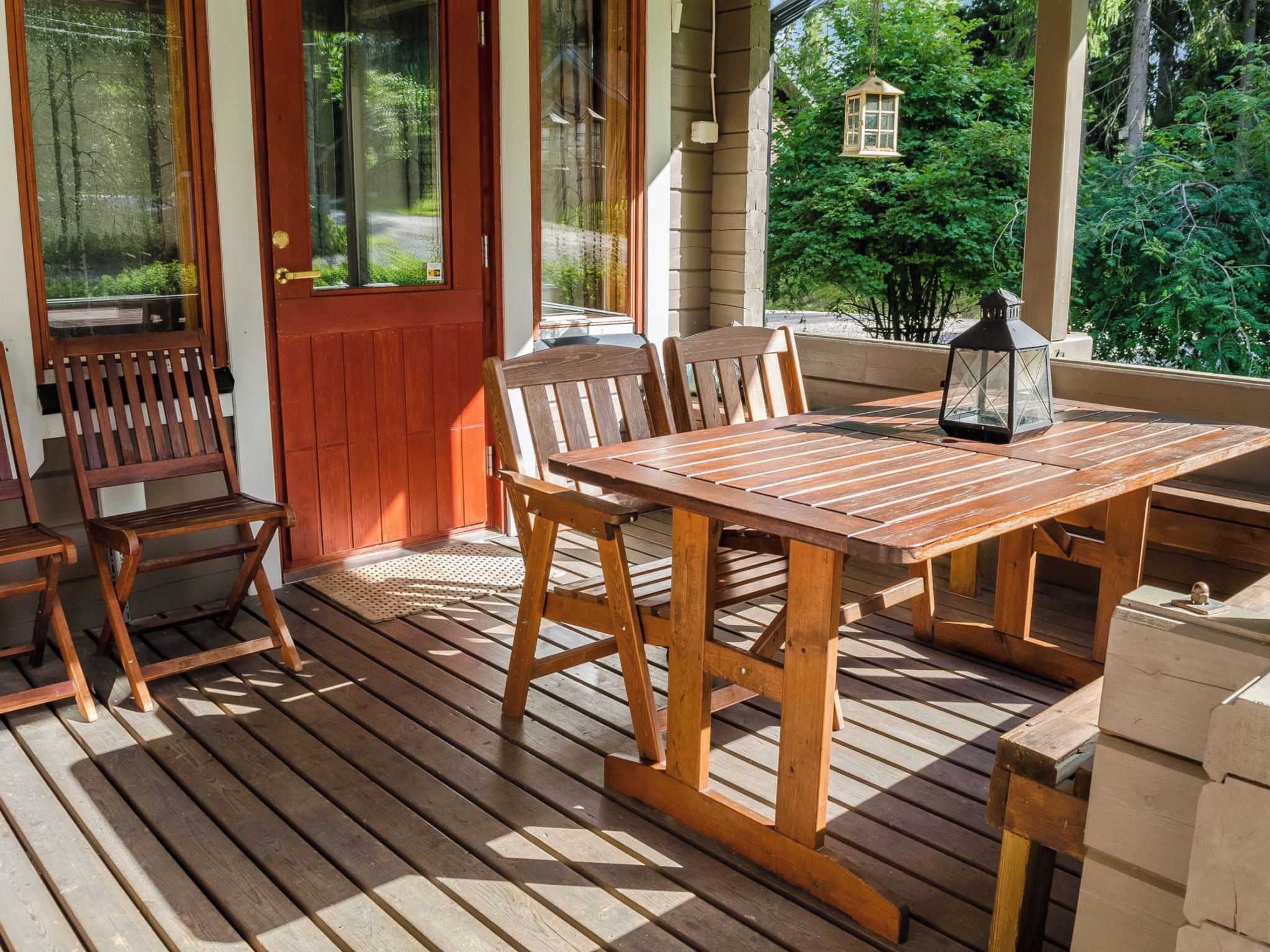 En træterrasse med et bord, stole, en dør og udsigt til træer.