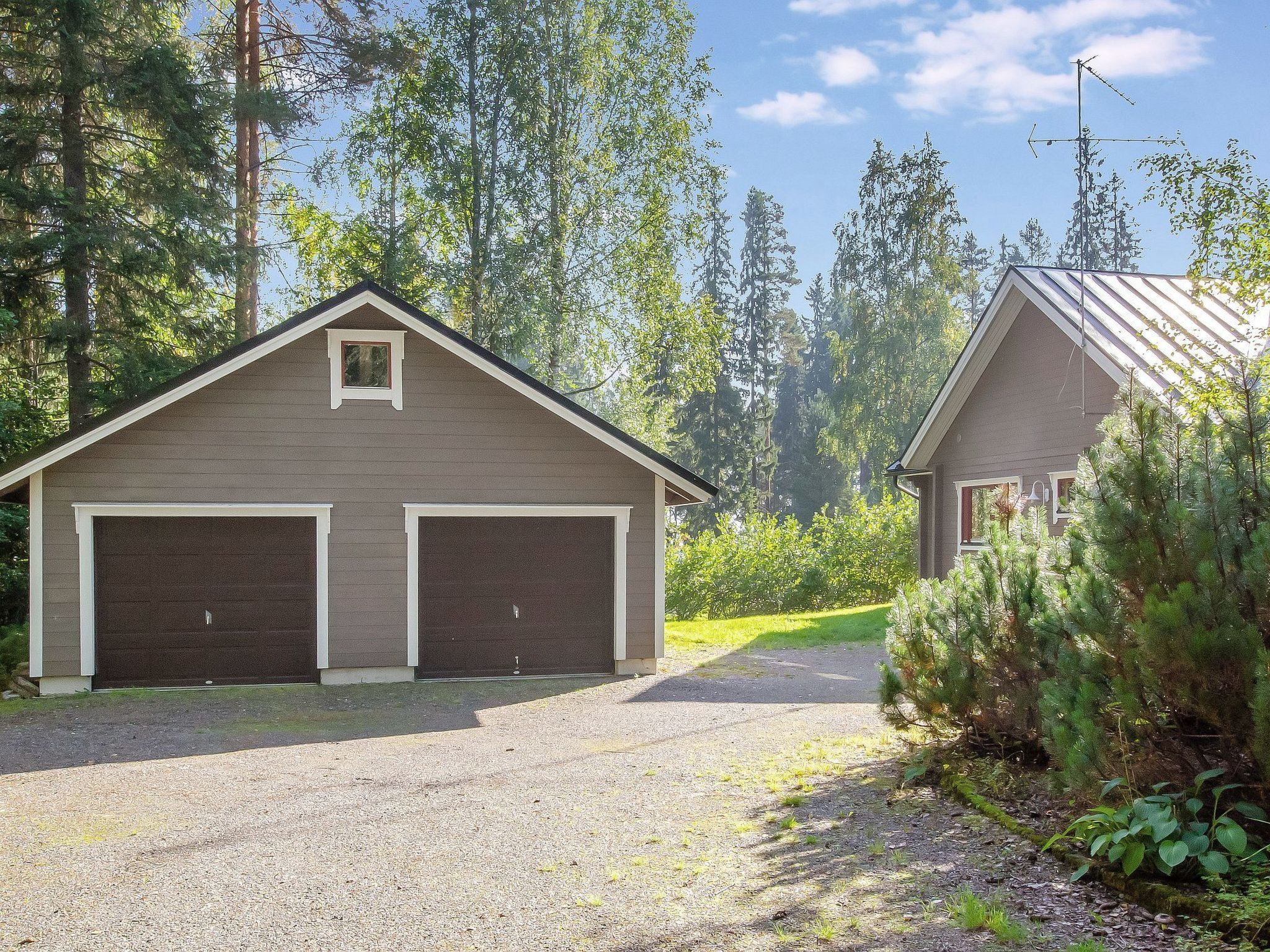 A two-car garage attached to a building with a gabled roof, surrounded by trees and a gravel driveway.