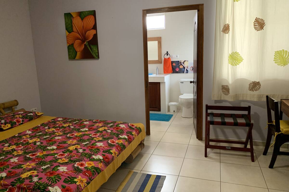 A bedroom with a bed, a chair, a bathroom visible through an open door, and a painting on the wall.