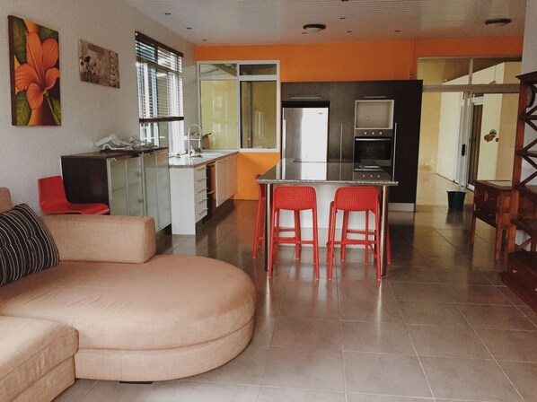 A modern living room with a curved sofa, a kitchen area with a dining table and red chairs, and a visible refrigerator. - City Home Tahiti (Papeete)