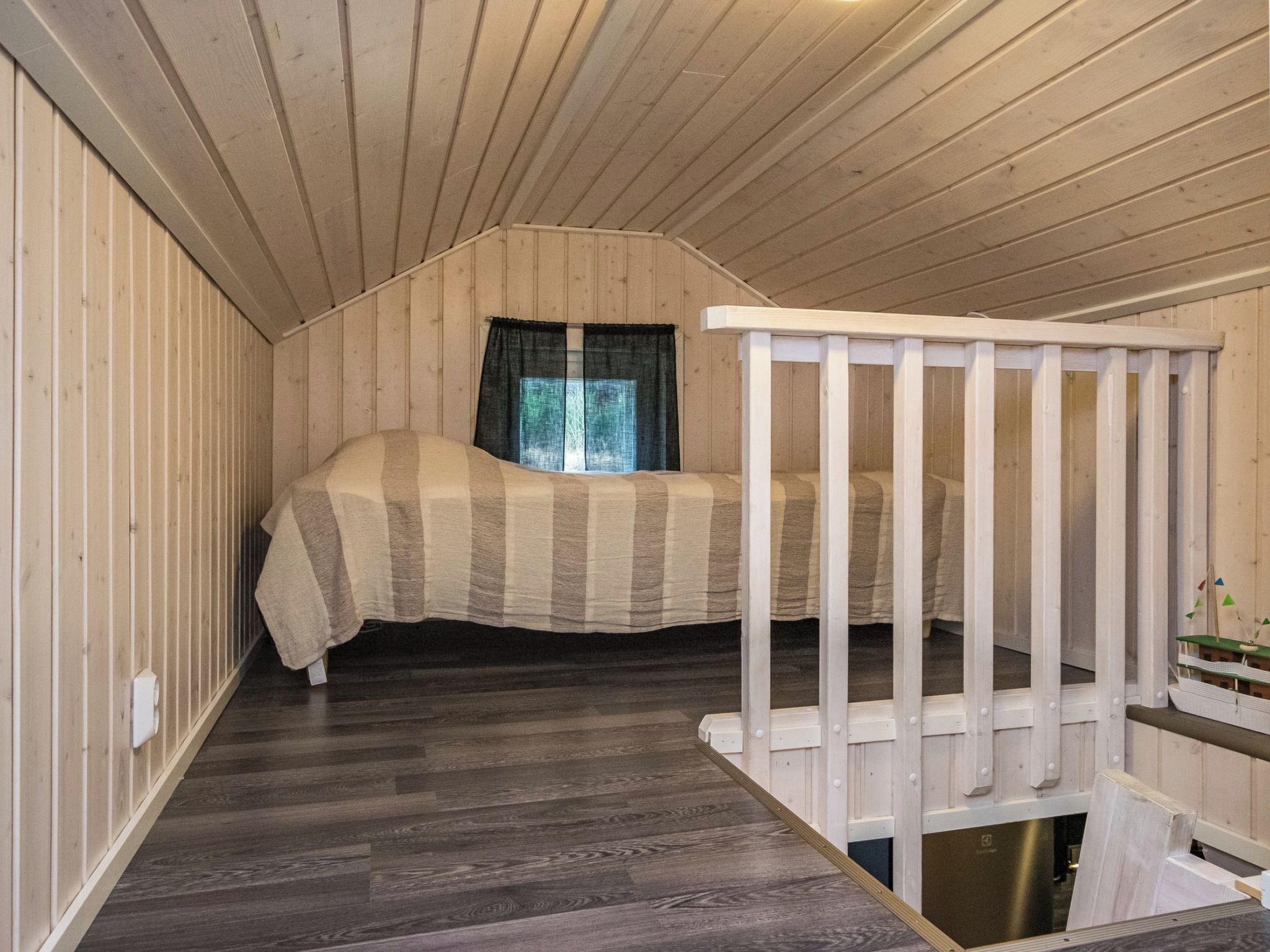 A bedroom with a bed, a window with curtains, a wooden floor, and a white railing.