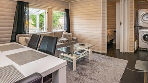 A modern living room with a sofa, coffee table, and a dining area. There is a view of a garden through the windows.