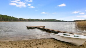 A boat docked on a sandy shore with a wooden pier extending into a calm lake, surrounded by dense forest.