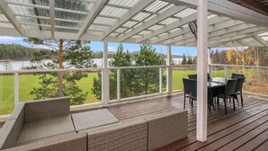 A covered outdoor deck with wicker furniture and a table, overlooking a lake and forest.
