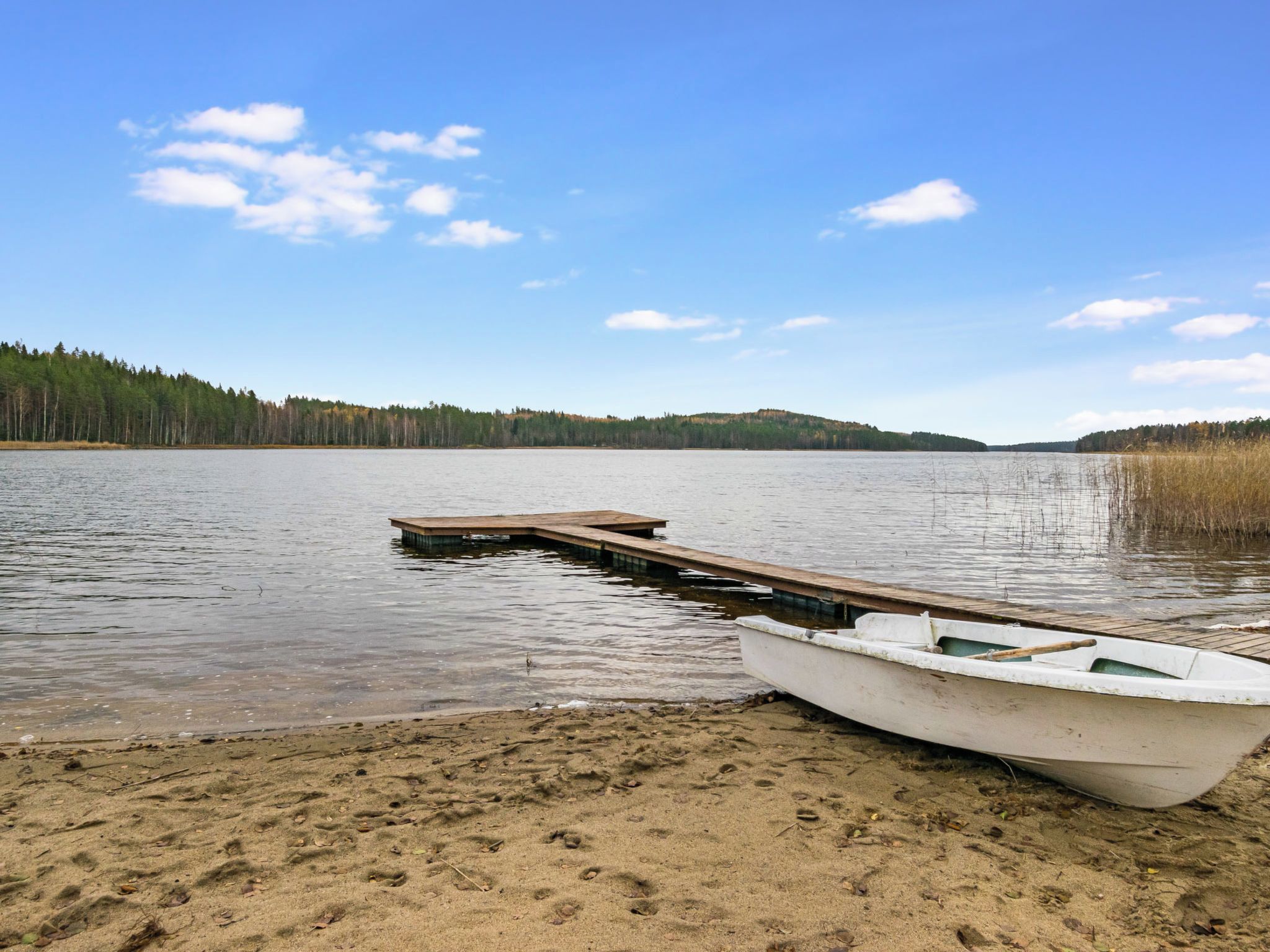 Un lac paisible, avec une jetée en bois qui s’avance sur l’eau, une petite barque à la rive et une colline boisée en arrière-plan.