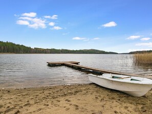 Un lac paisible, avec une jetée en bois qui s’avance sur l’eau, une petite barque à la rive et une colline boisée en arrière-plan.