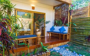 Um deck de madeira com um banco e uma mesa, cercado por plantas de bambu e uma cerca de bambu.