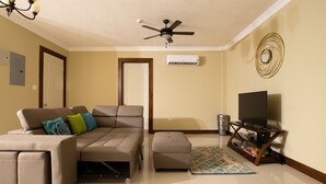 Una sala de estar con sofá modular, reposapiés, mueble para televisión, ventilador de techo y espejo decorativo.