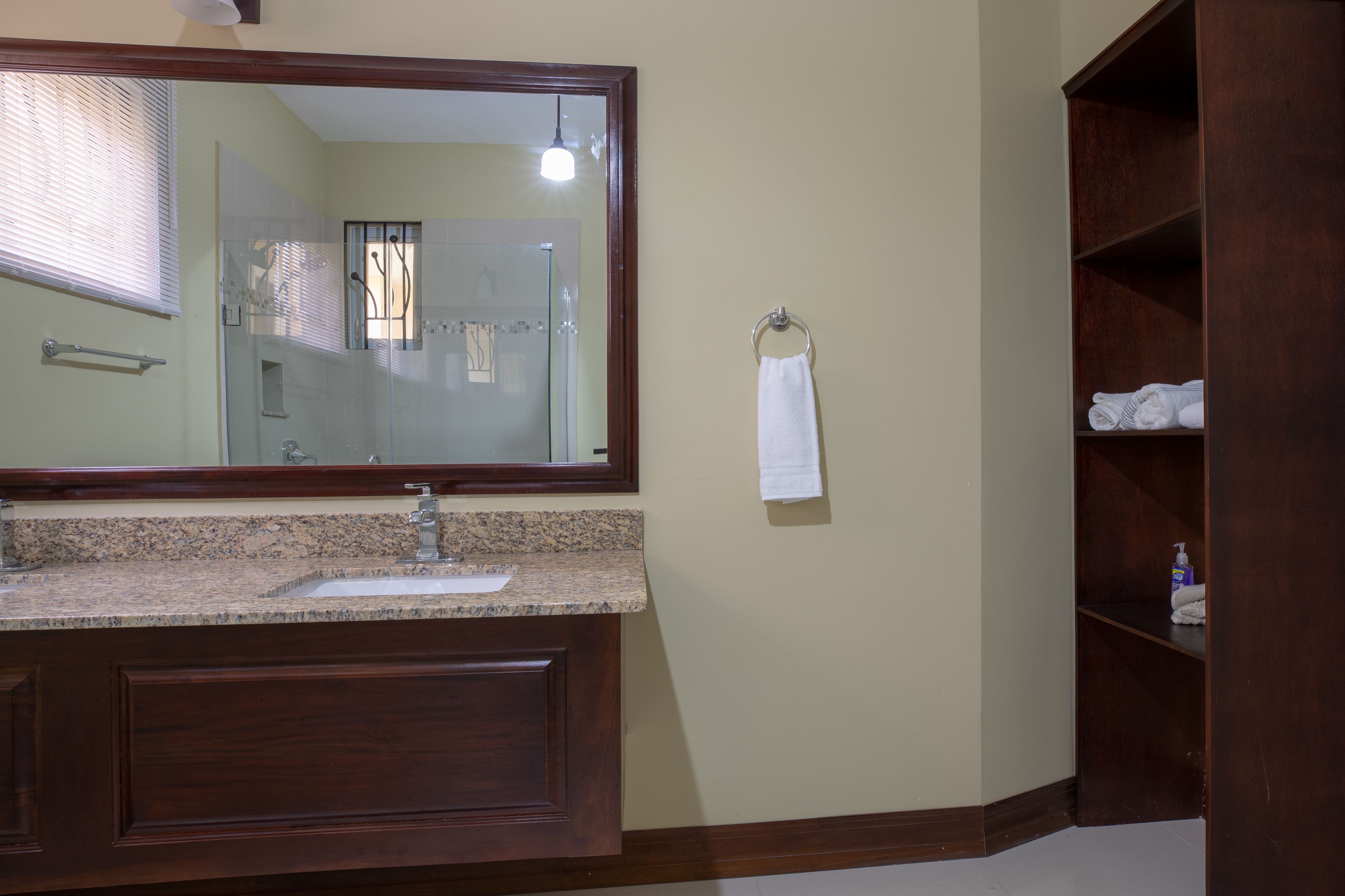A bathroom with a granite countertop sink, a large mirror, a towel rack, and a wooden shelving unit.