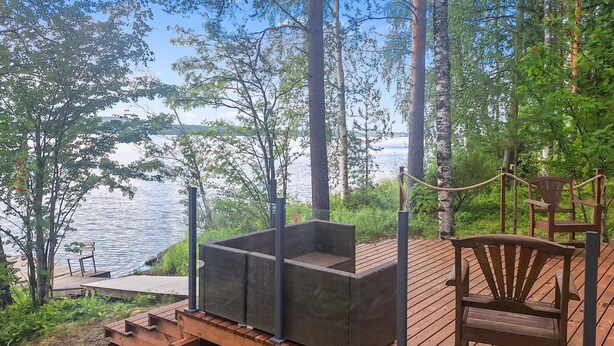 A wooden deck with a built-in seating area and a chair, overlooking a lake. - Honkaranta by Interhome (Karstula)
