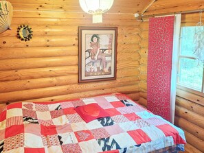Puinen mökkihuone, jossa on sänky, seinällä kehystetty kuva ja ikkuna, jossa on verhot.