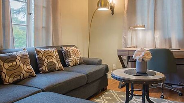 En moderne stue med en grå hjørnesofa, mønstrede puder, et rundt sofabord med en vase blomster, et skrivebord med en lampe og udsigt til grønne planter ude af vinduet.