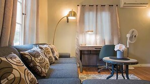 En hyggelig stue med en sofa, et skrivebord og en stol. Der er en lampe, en ventilator og et lille bord med en vase.