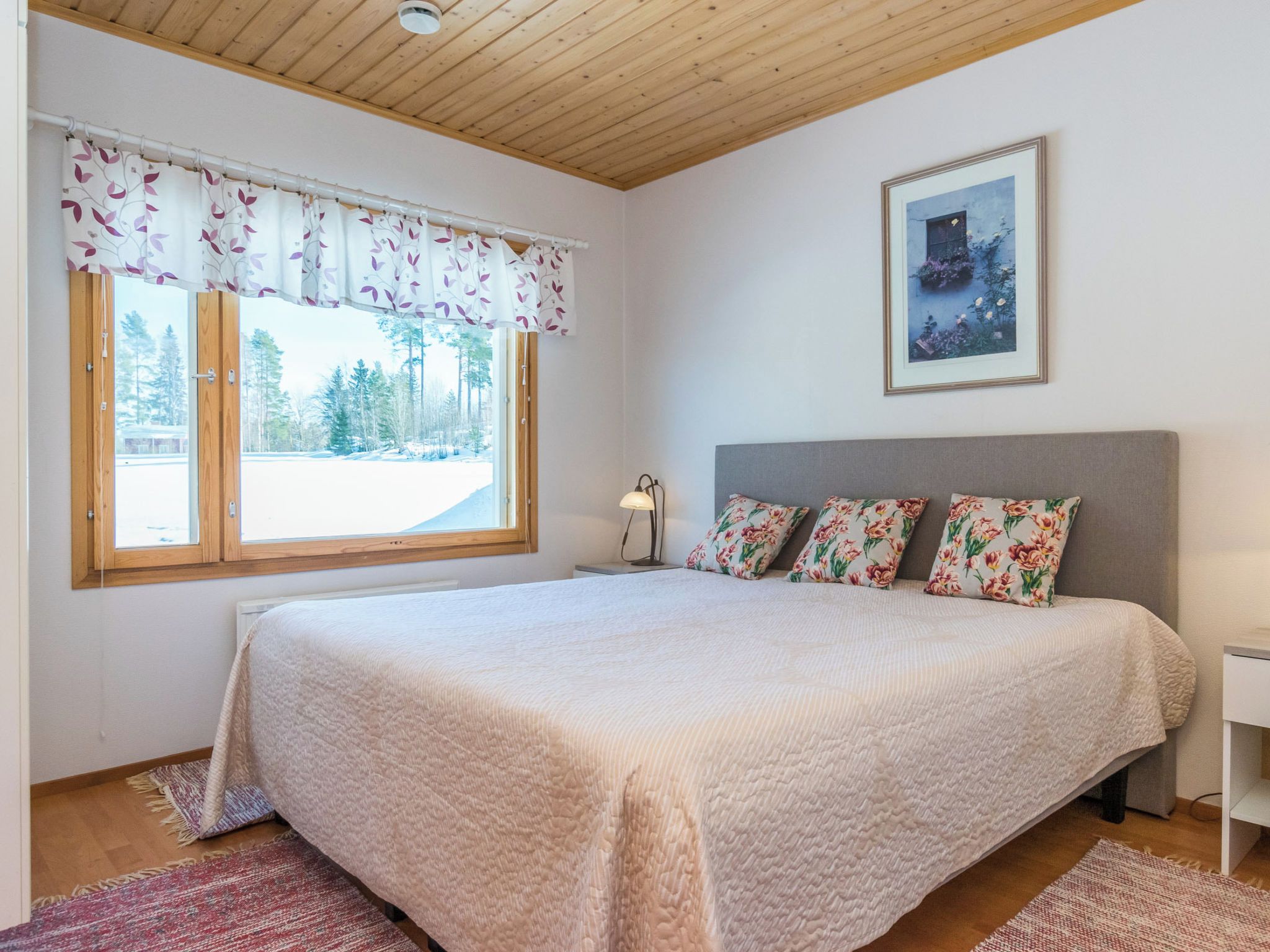 Un dormitorio con cama, ventana con cortinas, lámpara de noche y un cuadro en la pared.