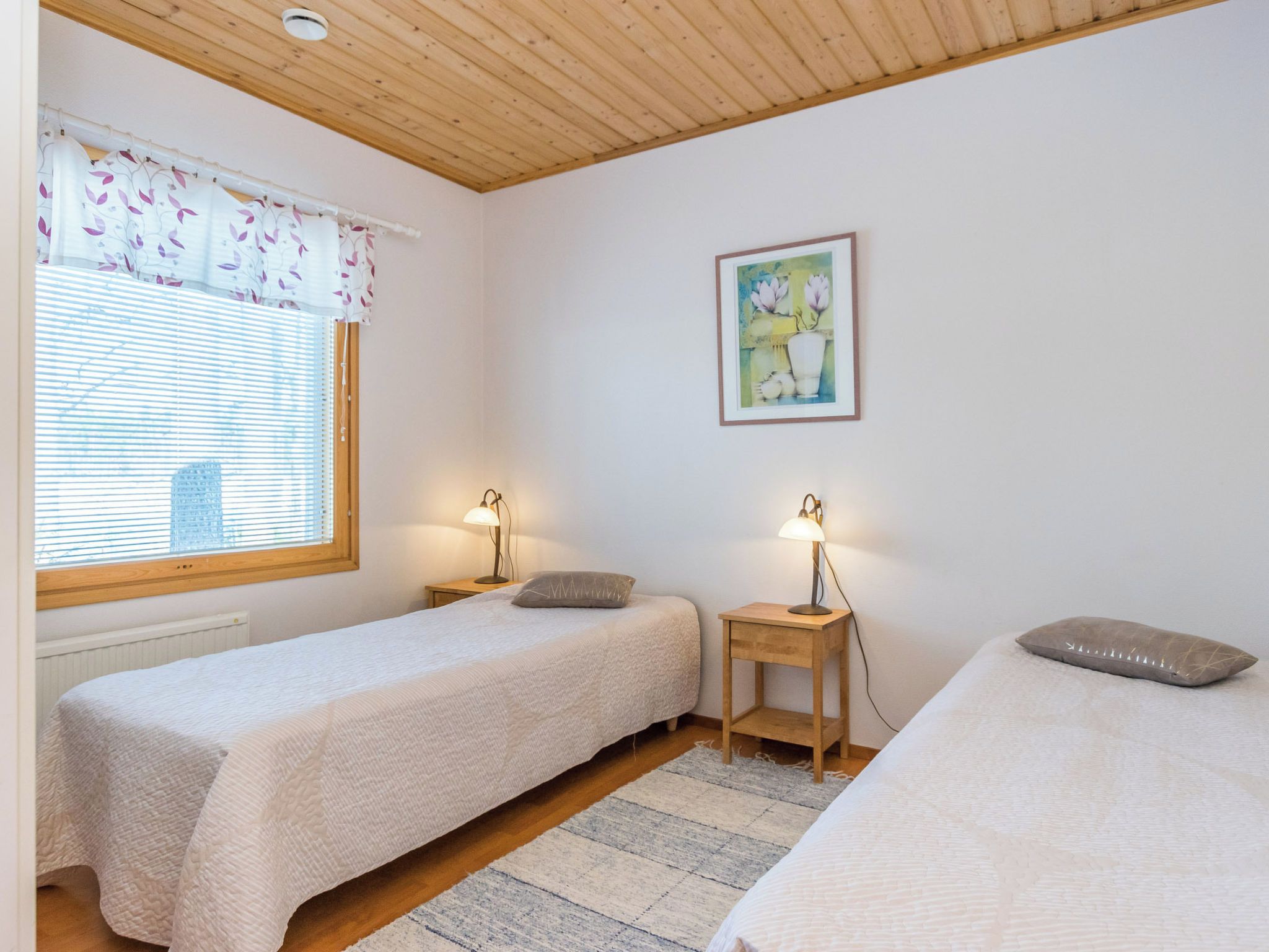 A bedroom with two beds, a window with blinds, a bedside table with a lamp, and a framed picture on the wall.