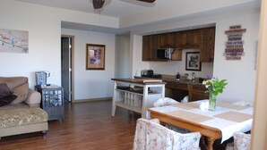 Amplia sala de estar con piso de madera, un sofá y una mesa de comedor preparada para cuatro comensales. Se ve una cocina con armarios de madera y un microondas al fondo.