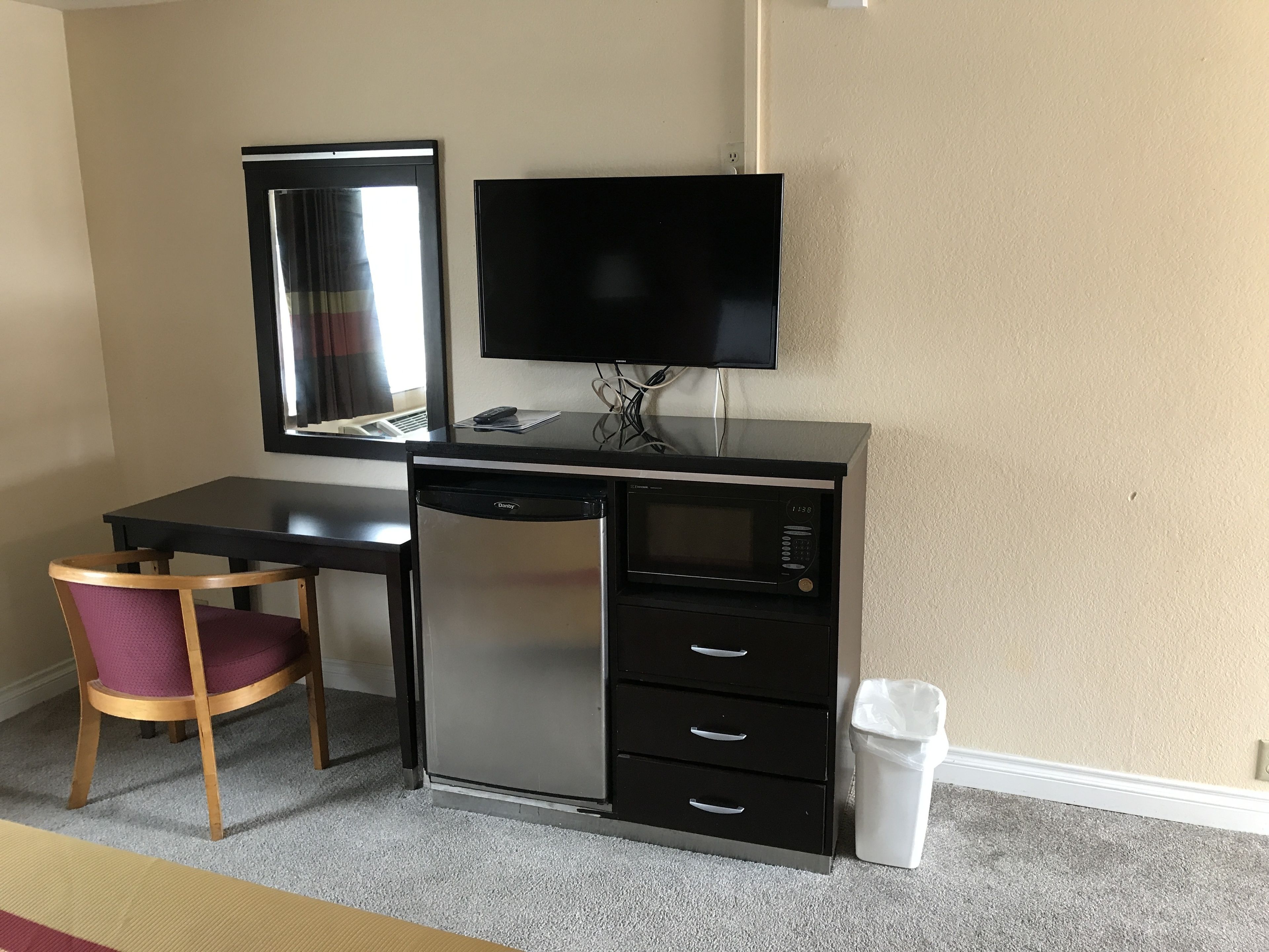 A compact hotel room with a kitchenette, a desk, a chair, a television, and a trash bin.