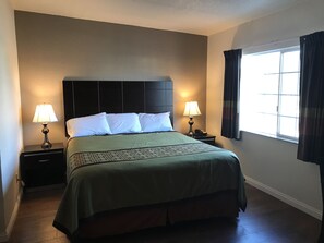 A hotel room with a bed, two bedside lamps, a telephone, a window with curtains, and a dark headboard. - Manhattan Inn & Suites - LAX (Manhattan Beach)