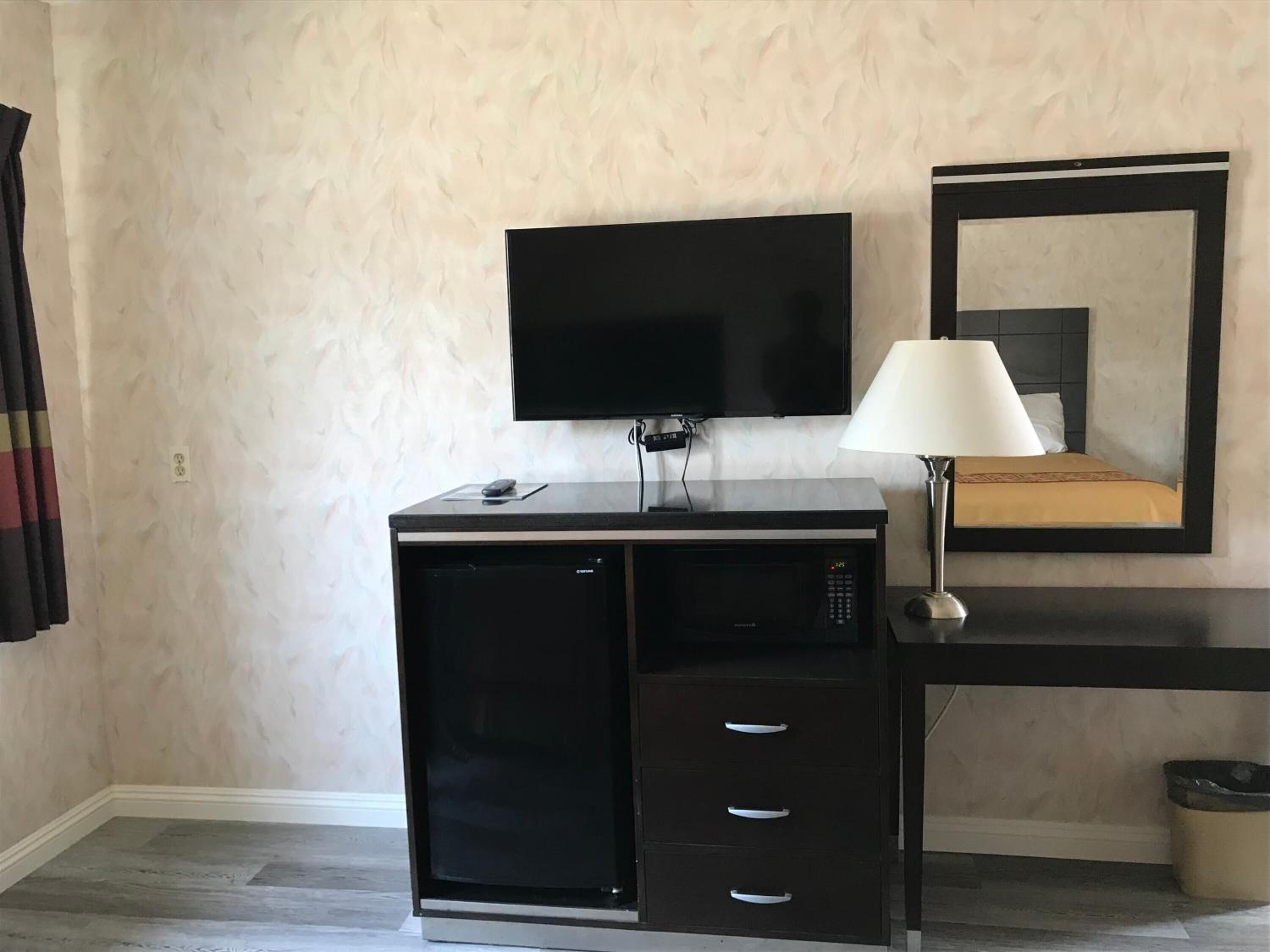 Quarto de hotel compacto com televisão de tela plana na parede, uma secretária pequena com luminária e uma cama com espelho em cima.