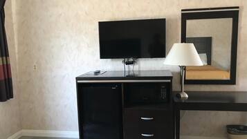 Quarto de hotel compacto com televisão de tela plana na parede, uma secretária pequena com luminária e uma cama com espelho em cima.