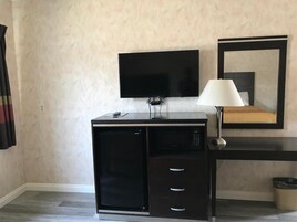 Quarto de hotel compacto com televisão de tela plana na parede, uma secretária pequena com luminária e uma cama com espelho em cima.