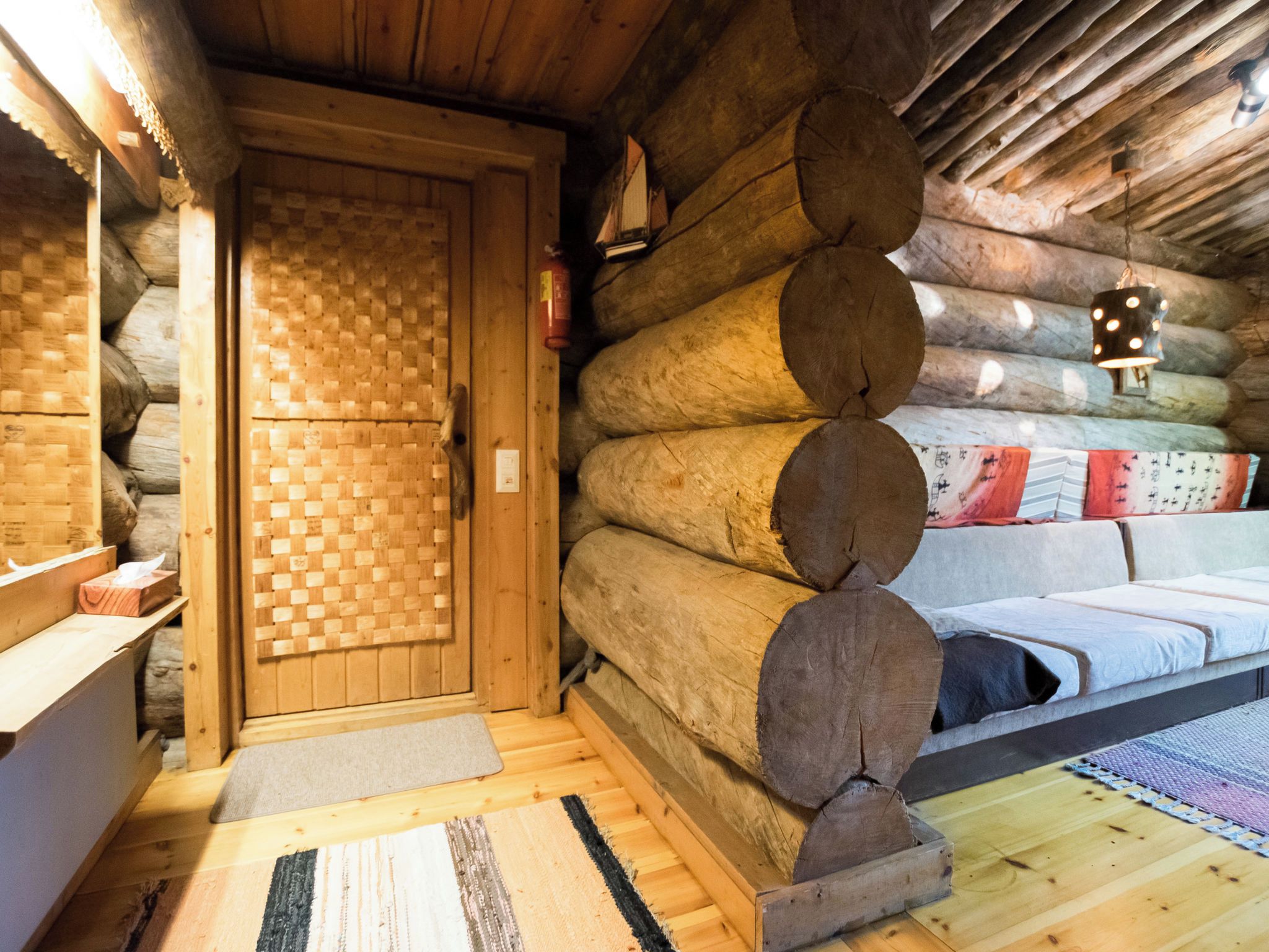 A log cabin interior with a woven door, a bed with a patterned blanket, and a hanging light fixture.