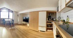 A compact kitchen with wooden cabinets, a dining area with chairs, and a television. - Ferienwohnung Nr. 4, für 1 bis 2 Personen, 46 qm, mit Dusche/WC, Wohn-/Schlafraum getrennt (Schönau am Königssee)